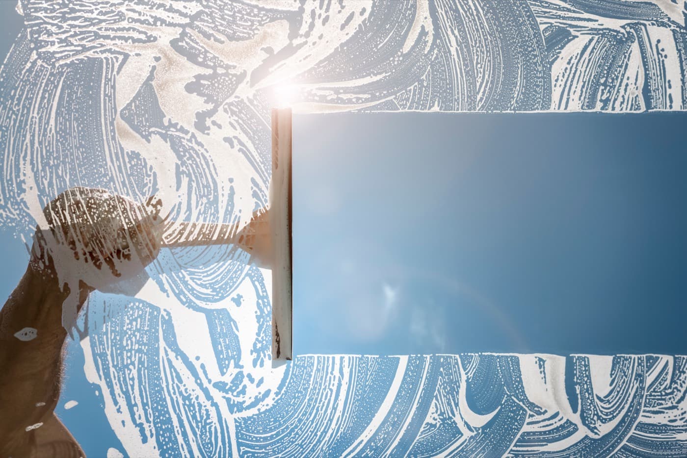 Hand using a squeegee to clean a soapy window and reveal blue sky